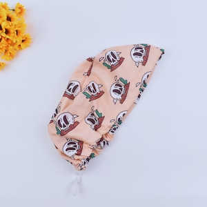 Puede incluir: Un gorro de quirófano de color melocotón con un patrón de calavera y huesos cruzados. El gorro tiene un cordón de ajuste y está hecho de una tela suave y cómoda.
