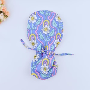 May include: A light purple scrub cap with a floral pattern of smiling daisies, yellow rainbows, and green leaves. The cap has a drawstring closure. Ideal for medical or beauty professionals. The cap's design is reminiscent of the 1970s.