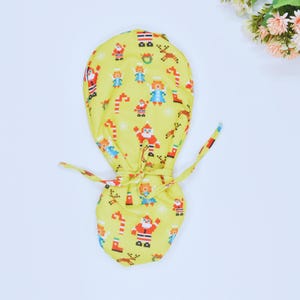 May include: A yellow scrub cap with a Christmas-themed pattern. The cap features pixelated images of Santa Claus, elves, reindeer, and candy canes. The cap has a drawstring closure and is made of a soft fabric.