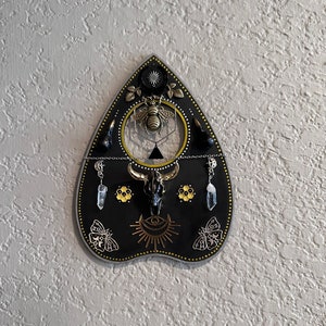 May include: Black wooden Ouija board with gold accents, featuring a bee, skull, and crescent moon design. The board has a hanging crystal charm and a chain.