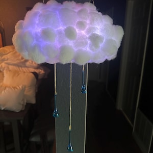 Könnte beinhalten: Eine wolkenförmige dekorative Leuchte. Die Wolke besteht aus weißem Material mit interner lila und blauer Beleuchtung. Goldketten hängen die Wolke auf, mit blauen tropfenförmigen Ornamenten darunter.
