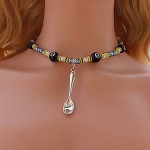 May include: A black and yellow beaded choker necklace with a silver spoon charm. The beads are round and have a black and yellow striped pattern. The spoon is hanging from the center of the necklace.