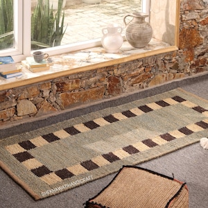May include: A rectangular woven runner rug featuring a central sage green panel bordered by a repeating pattern of alternating dark brown and beige squares. The text SHEKHAWATIHANDICRAFT is printed in white on the lower left corner of the rug.