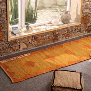 Peut inclure: Un long tapis rectangulaire avec un motif géométrique dans les tons orange et jaune. Le tapis est posé sur une surface grise, avec un pouf tissé au premier plan. Le texte "SHEKHAWATIHANDICRAFT" est visible sur le tapis.