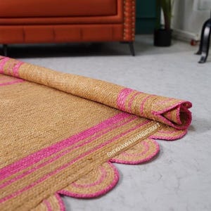 Op de afbeelding: Een natuurlijk beige jute vloerkleed met felroze strepen en een geschulpte rand. Het kleed is gedeeltelijk opgerold, waardoor de geweven textuur zichtbaar is. De tekst SHEKHAWATIHANDICRAFT is in het wit op het kleed gedrukt.