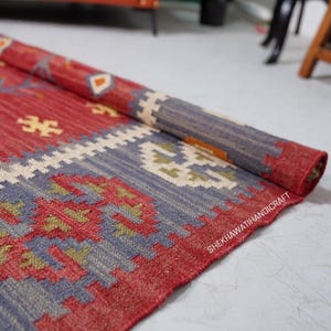 May include: Rolled-up red, blue, and cream-colored rug with geometric patterns. The rug features a mix of colors and designs, with a prominent red base and detailed borders. The rug is made of woven material.