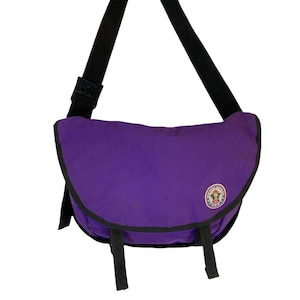 May include: A purple messenger bag with a black adjustable shoulder strap and black trim. The bag has a circular patch with text and a graphic. Two black straps are visible on the bottom.