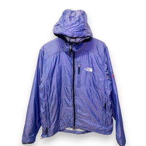 May include: A periwinkle blue hooded jacket with a full-length black zip. The jacket features a small white logo on the chest and a red and black logo on the sleeve. It is made of a lightweight, shiny material.