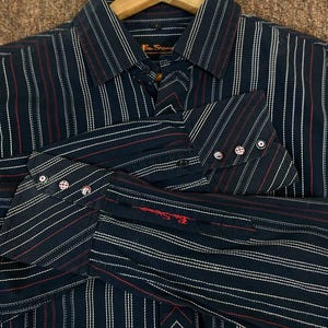 Op de afbeelding: Een marineblauw overhemd met verticale strepen in wit en rood. Het overhemd heeft een klassieke kraag, lange mouwen en het Ben Sherman-logo, rood geborduurd op de manchet. Het hangt aan een houten kleerhanger.