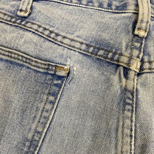 May include: Close-up of light blue denim jeans, showing the back pocket and waistband. The jeans have a faded wash and visible stitching details. There is a small tear near the pocket.