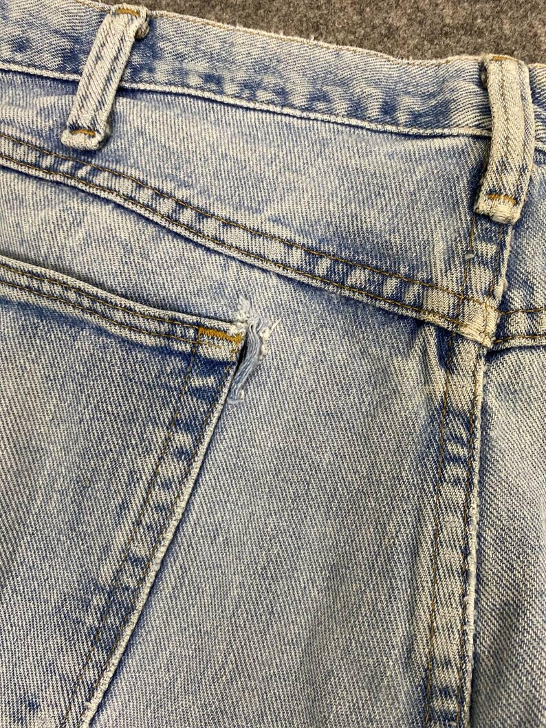 May include: Close-up of light blue denim jeans, showing the back pocket and waistband. The jeans have a faded wash and visible stitching details. There is a small tear near the pocket.