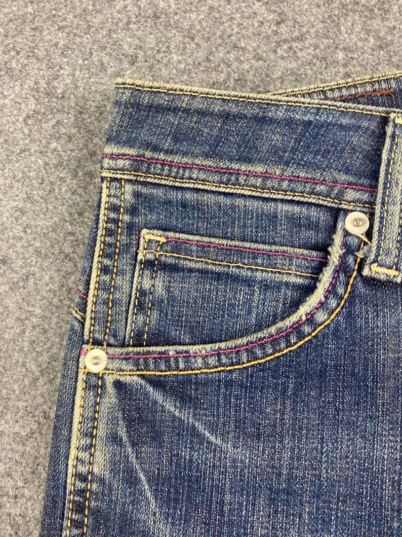 Puede incluir: Primer plano del bolsillo de unos vaqueros de mezclilla azul con costuras amarillas y rosas. Los vaqueros tienen cierre de bot&oacute;n y un bolsillo peque&ntilde;o. La mezclilla tiene un aspecto descolorido y desgastado.