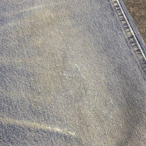 May include: Close-up of light blue denim fabric with distressed areas. The denim shows signs of wear and tear, with faded patches and a small tear. The texture of the fabric is visible, highlighting the weave of the denim.