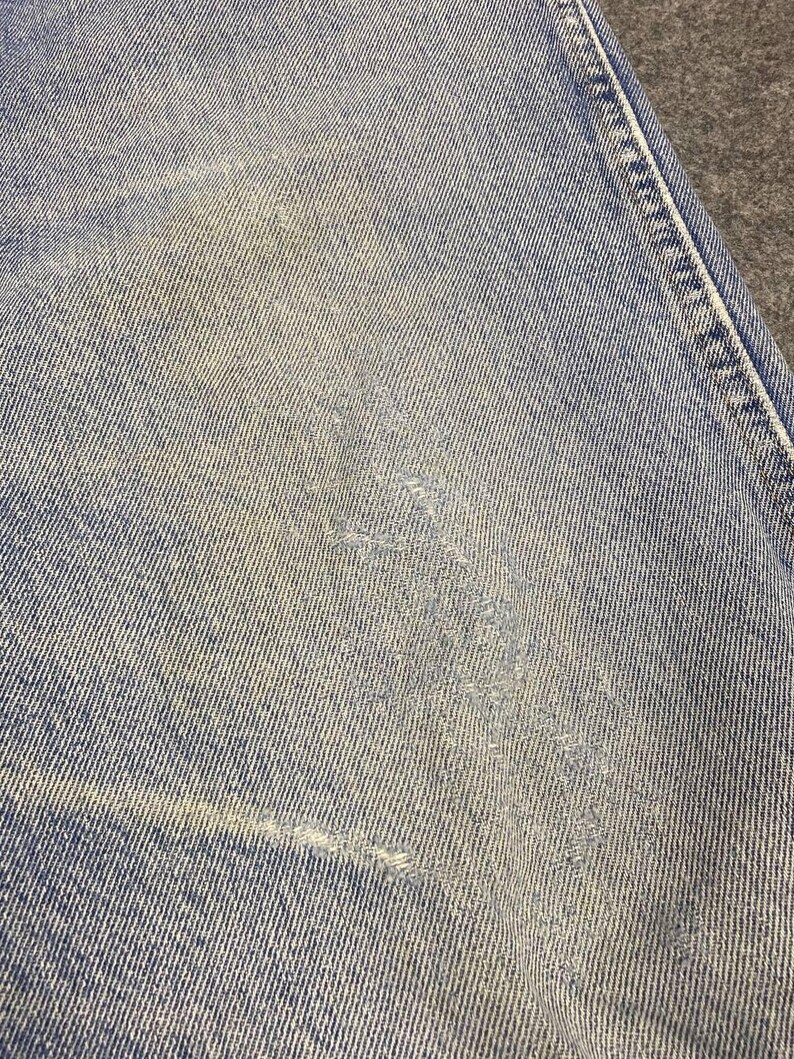 May include: Close-up of light blue denim fabric with distressed areas. The denim shows signs of wear and tear, with faded patches and a small tear. The texture of the fabric is visible, highlighting the weave of the denim.