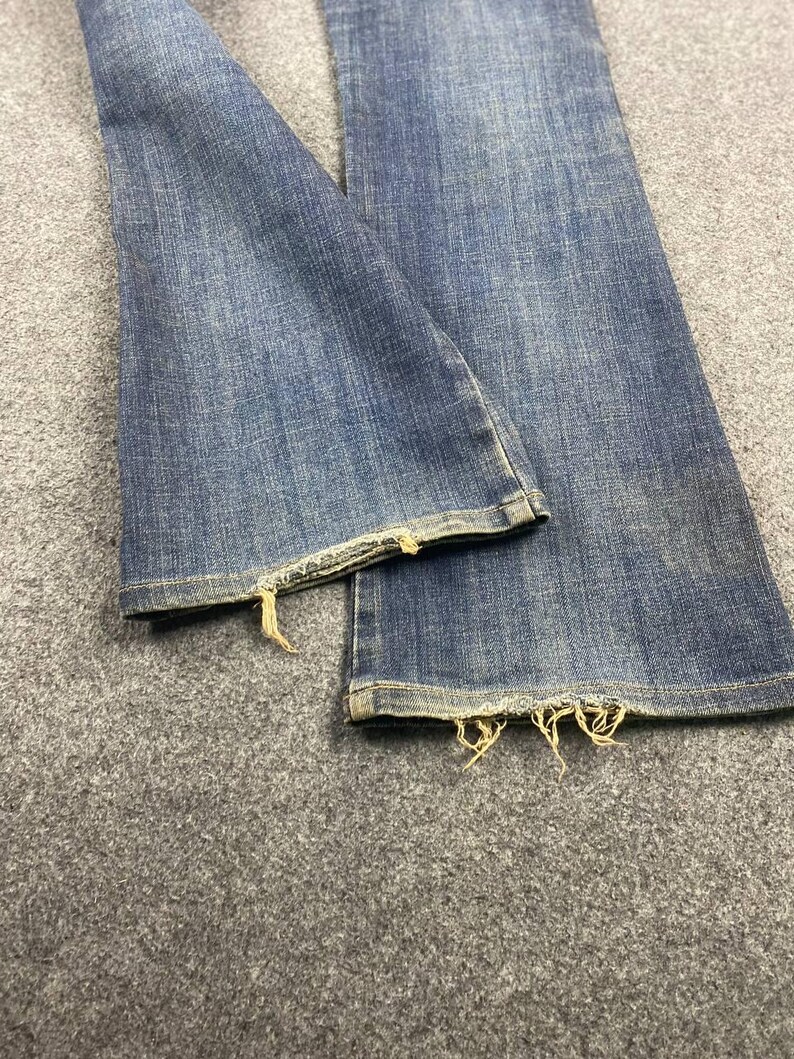 Puede incluir: Primer plano de vaqueros de mezclilla azules con dobladillos deshilachados. Los vaqueros tienen un dise&ntilde;o cl&aacute;sico de cinco bolsillos y un lavado medio. Los bordes deshilachados a&ntilde;aden un aspecto desgastado a los pantalones vaqueros.