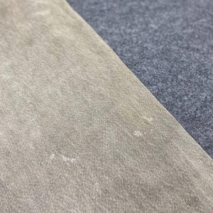May include: Close-up of a section of tan-colored denim fabric. The fabric has a textured appearance with visible vertical lines and subtle variations in color. The denim is angled against a contrasting gray fabric.