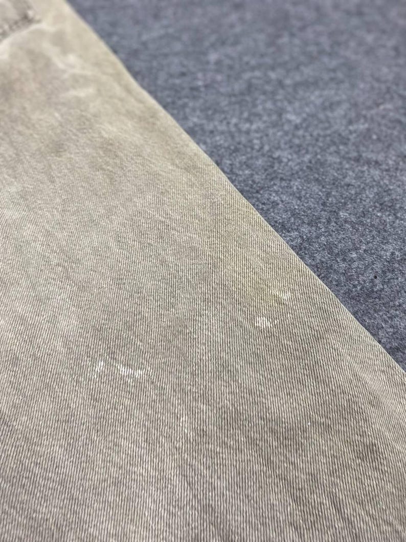May include: Close-up of a section of tan-colored denim fabric. The fabric has a textured appearance with visible vertical lines and subtle variations in color. The denim is angled against a contrasting gray fabric.