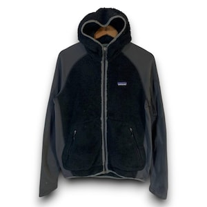 Op de afbeelding: Een zwarte fleecejas met capuchon en grijze mouwen en afwerking. De jas heeft een ritssluiting over de hele lengte, twee voorzakken en een kleine logopatch op de borst. De capuchon is gevoerd met een zacht, donzig materiaal.