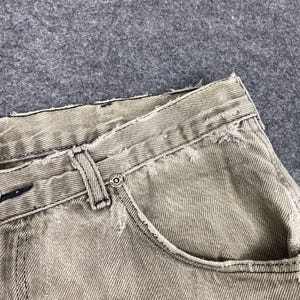 May include: Close-up of distressed, light gray denim jeans. The jeans feature a classic five-pocket design with a button closure and belt loops. Frayed edges and subtle fading add to the vintage aesthetic. The fabric appears to be a medium weight.