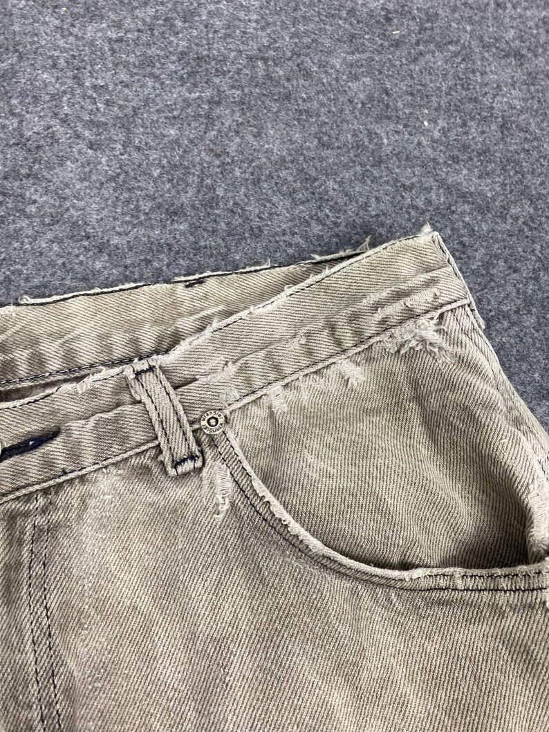 May include: Close-up of distressed, light gray denim jeans. The jeans feature a classic five-pocket design with a button closure and belt loops. Frayed edges and subtle fading add to the vintage aesthetic. The fabric appears to be a medium weight.