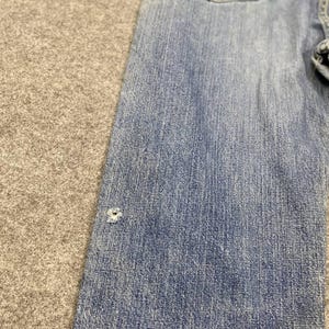 May include: Close-up of a section of light blue denim jeans. The jeans have a classic five-pocket design and a faded appearance. A small hole is visible on the front of the jeans. The fabric has a textured appearance.