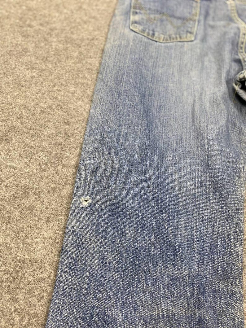 May include: Close-up of a section of light blue denim jeans. The jeans have a classic five-pocket design and a faded appearance. A small hole is visible on the front of the jeans. The fabric has a textured appearance.