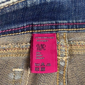 Puede incluir: Primer plano de unos vaqueros de mezclilla con una etiqueta rosa de Wrangler. La etiqueta presenta el logotipo de Wrangler e instrucciones de lavado. Los vaqueros tienen costuras rosas y un lavado de mezclilla cl&aacute;sico.