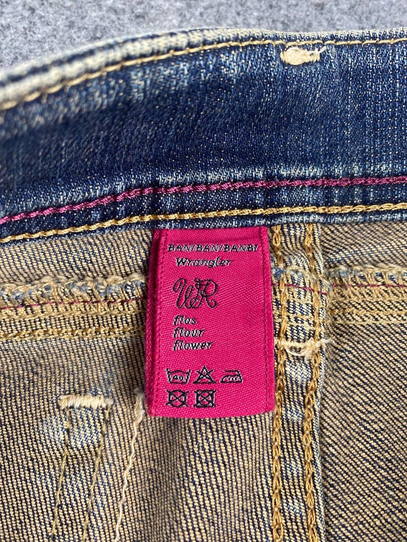 Puede incluir: Primer plano de unos vaqueros de mezclilla con una etiqueta rosa de Wrangler. La etiqueta presenta el logotipo de Wrangler e instrucciones de lavado. Los vaqueros tienen costuras rosas y un lavado de mezclilla cl&aacute;sico.