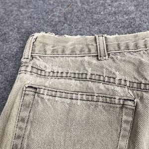 May include: Close-up of the back of light tan denim jeans. The jeans have a distressed look with frayed edges and black stitching. The back pocket is visible, along with the belt loops and waistband.