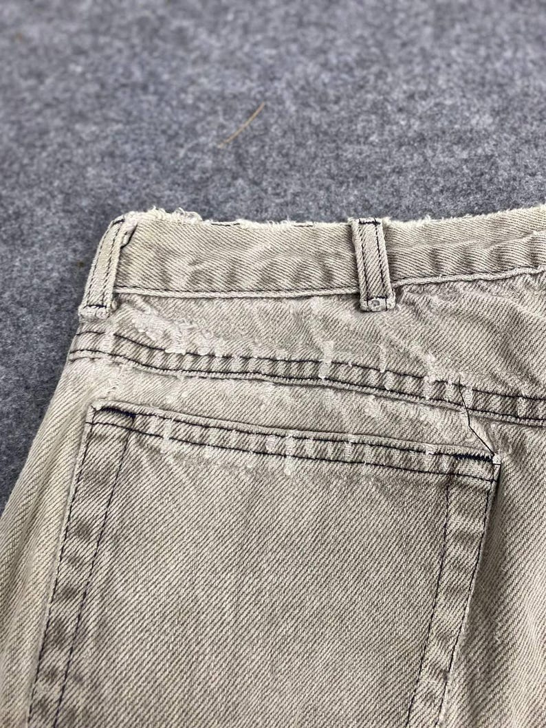 May include: Close-up of the back of light tan denim jeans. The jeans have a distressed look with frayed edges and black stitching. The back pocket is visible, along with the belt loops and waistband.
