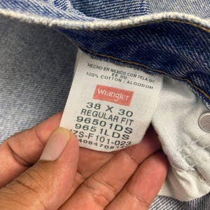 May include: Close-up of a pair of blue denim jeans with a Wrangler label. The label reads "38 X 30 REGULAR FIT" and "100% COTTON". The jeans are made in Mexico.