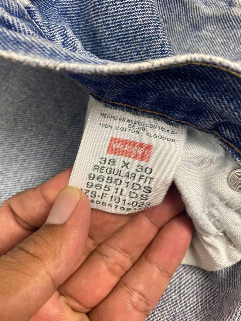 May include: Close-up of a pair of blue denim jeans with a Wrangler label. The label reads "38 X 30 REGULAR FIT" and "100% COTTON". The jeans are made in Mexico.