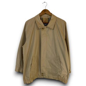 May include: A beige windbreaker jacket featuring a classic collar, a front zipper closure concealed by a placket, and two side pockets. The interior lining displays a tan, black, and red plaid pattern. The jacket has long sleeves and an elastic hem.