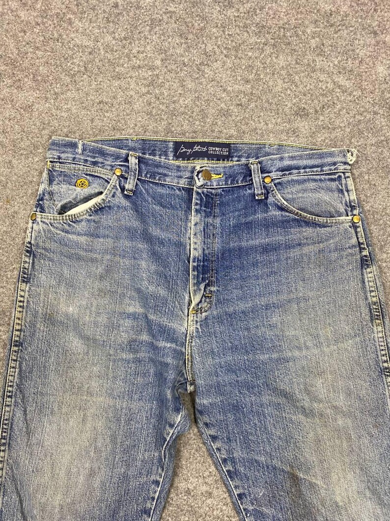 May include: Blue denim jeans with a classic five-pocket design. The jeans feature a button and zipper closure, belt loops, and brass-colored rivets. The "Cowboy Cut Collection" label is visible on the waistband. The jeans have a faded wash and a straight leg.