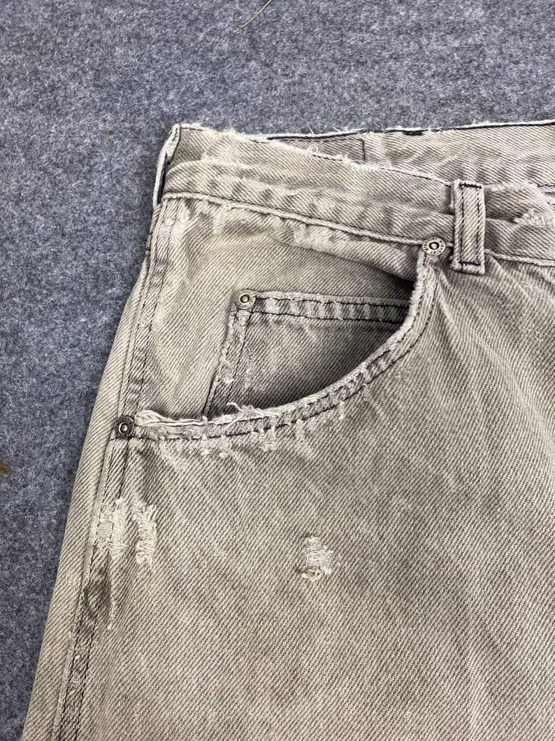 May include: Close-up of light gray denim jeans with distressed details. The jeans feature a classic five-pocket design, with visible wear and tear around the pockets and seams. The fabric has a faded appearance, suggesting a worn-in look.
