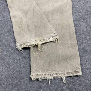May include: Close-up of a pair of light gray denim jeans with distressed, frayed hems. The jeans are folded, showing the texture and wear of the fabric. The color is a muted, neutral tone, suggesting a vintage or worn aesthetic.