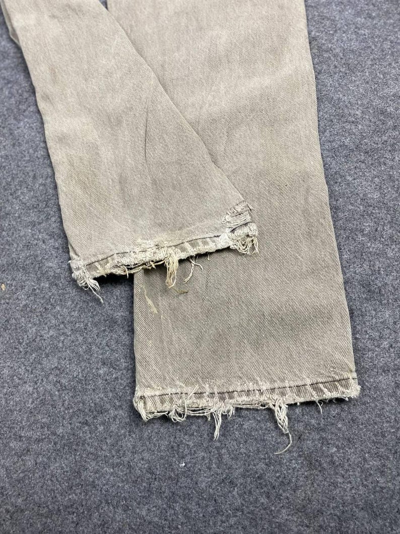May include: Close-up of a pair of light gray denim jeans with distressed, frayed hems. The jeans are folded, showing the texture and wear of the fabric. The color is a muted, neutral tone, suggesting a vintage or worn aesthetic.