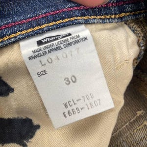 Puede incluir: Primer plano del interior de unos vaqueros azules. Una etiqueta blanca muestra el logotipo "Wrangler" y texto. La etiqueta indica la talla 30. Los vaqueros tienen costuras rosas y doradas.