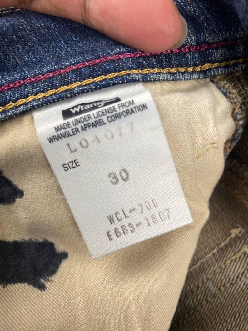 Puede incluir: Primer plano del interior de unos vaqueros azules. Una etiqueta blanca muestra el logotipo "Wrangler" y texto. La etiqueta indica la talla 30. Los vaqueros tienen costuras rosas y doradas.