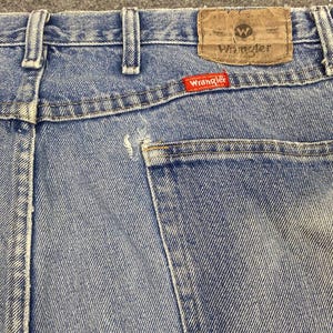 May include: Close-up of the back of light blue denim jeans. The jeans feature a brown leather patch with the Wrangler logo and a red Wrangler tag. The back pocket is visible, and the stitching is white.