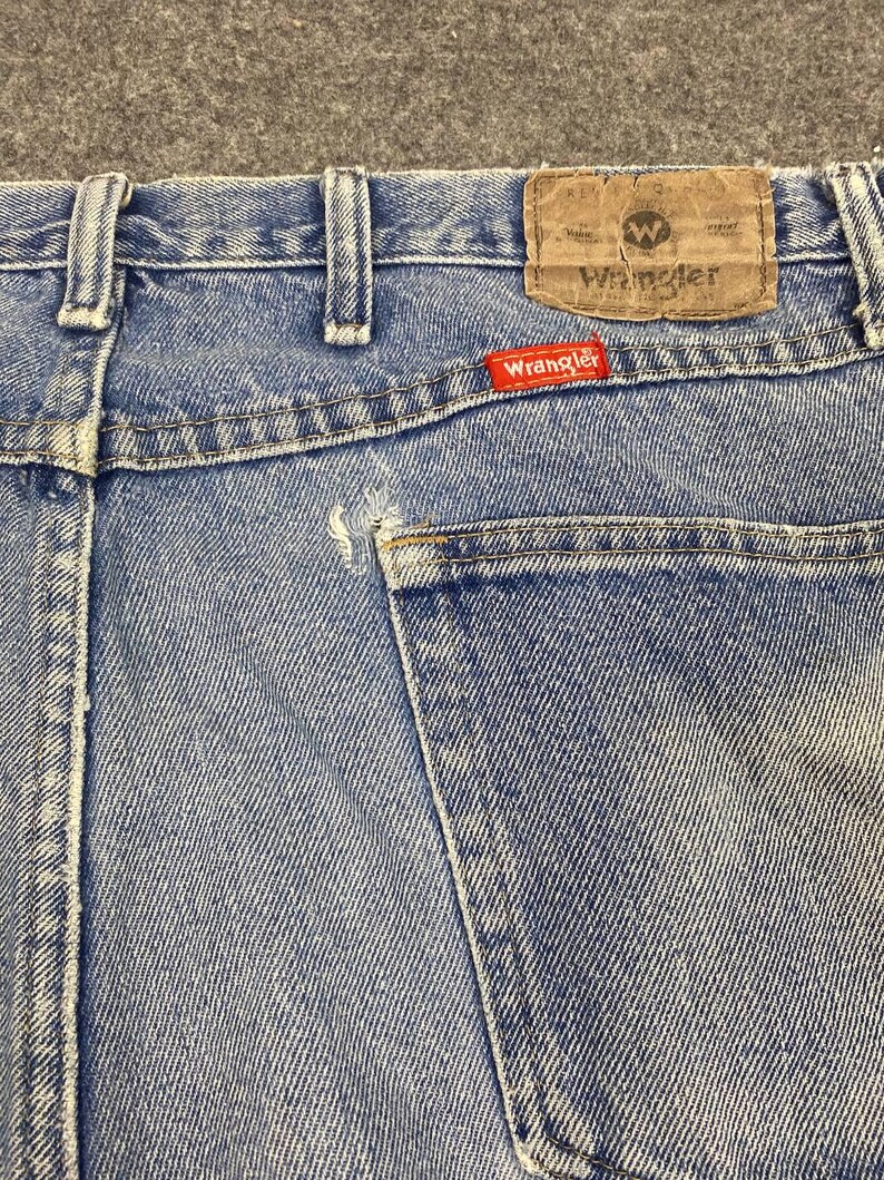 May include: Close-up of the back of light blue denim jeans. The jeans feature a brown leather patch with the Wrangler logo and a red Wrangler tag. The back pocket is visible, and the stitching is white.