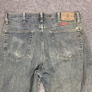 Może przedstawiać: Spodnie dżinsowe z wyblakłego niebieskiego denimu z klasycznym pięciokieszeniowym designem. Etykieta marki Wrangler jest widoczna na tylnej kieszeni. Dżinsy noszą ślady zużycia, co dodaje im uroku vintage.