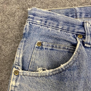 May include: Close-up of light blue denim jeans. The jeans have a classic five-pocket design with brass-colored rivets and a distressed pocket edge. The fabric shows signs of wear.