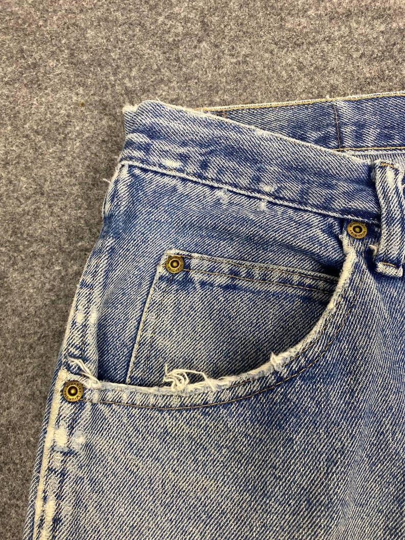 May include: Close-up of light blue denim jeans. The jeans have a classic five-pocket design with brass-colored rivets and a distressed pocket edge. The fabric shows signs of wear.