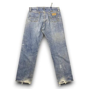 May include: Faded blue denim jeans with a classic five-pocket design. The jeans feature a worn look with distressed details at the hem and a leather patch on the back pocket. The Wrangler logo is visible.