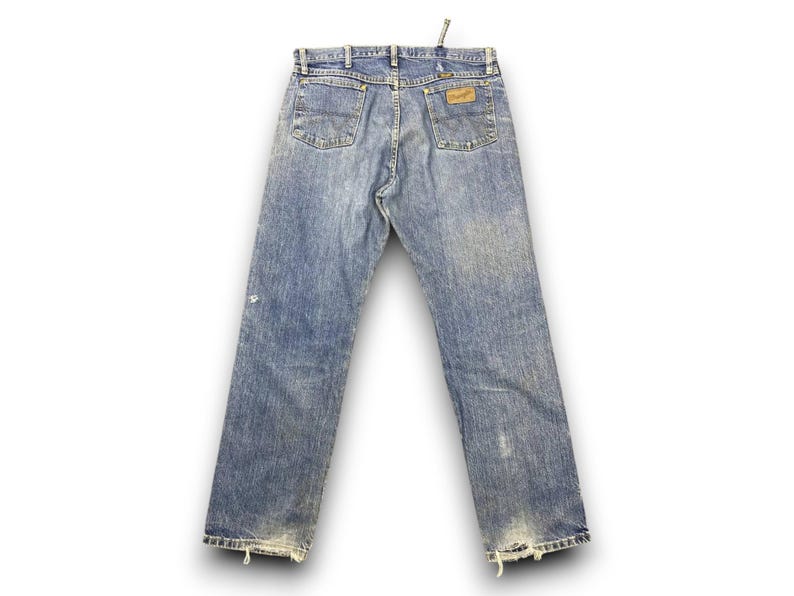 May include: Faded blue denim jeans with a classic five-pocket design. The jeans feature a worn look with distressed details at the hem and a leather patch on the back pocket. The Wrangler logo is visible.