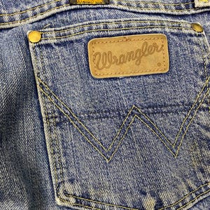 May include: Close-up of blue denim jeans featuring a back pocket with the "Wrangler" logo on a brown leather patch. The pocket is stitched with yellow thread, and the jeans show signs of wear.