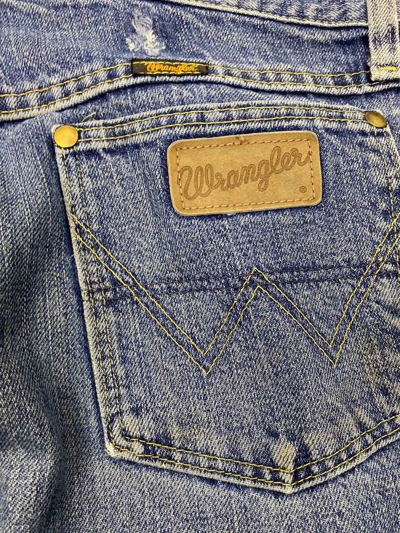 May include: Close-up of blue denim jeans featuring a back pocket with the "Wrangler" logo on a brown leather patch. The pocket is stitched with yellow thread, and the jeans show signs of wear.