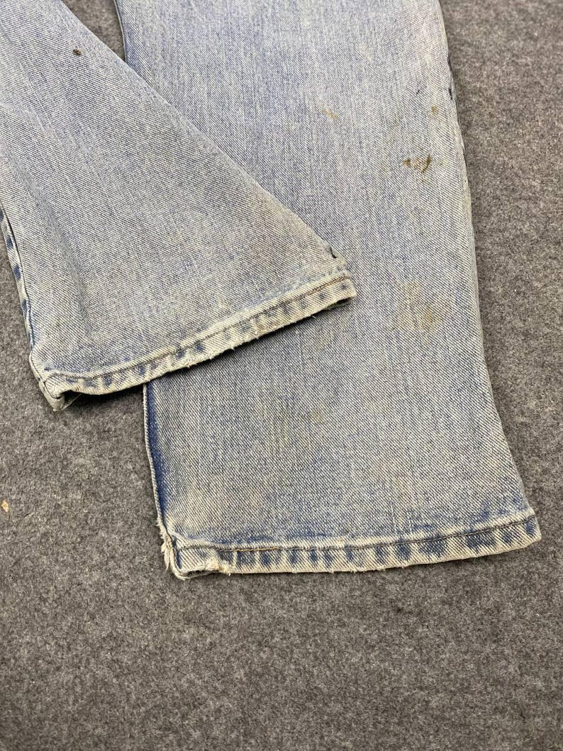 K&ouml;nnte beinhalten: Nahaufnahme einer hellblauen Jeans. Die Jeans weist Gebrauchsspuren auf, mit sichtbaren Flecken und Ausfransungen am Saum. Der Stoff hat ein verwaschenes Aussehen, was auf Alter oder Waschen hindeutet. Das Bild hebt die Textur und Details des Jeansstoffs hervor.