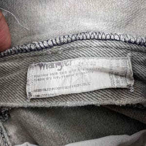 May include: Close-up of gray denim jeans with a white label. The label reads "Wrangler" and includes size information. The jeans have visible stitching and are made of 100% cotton.